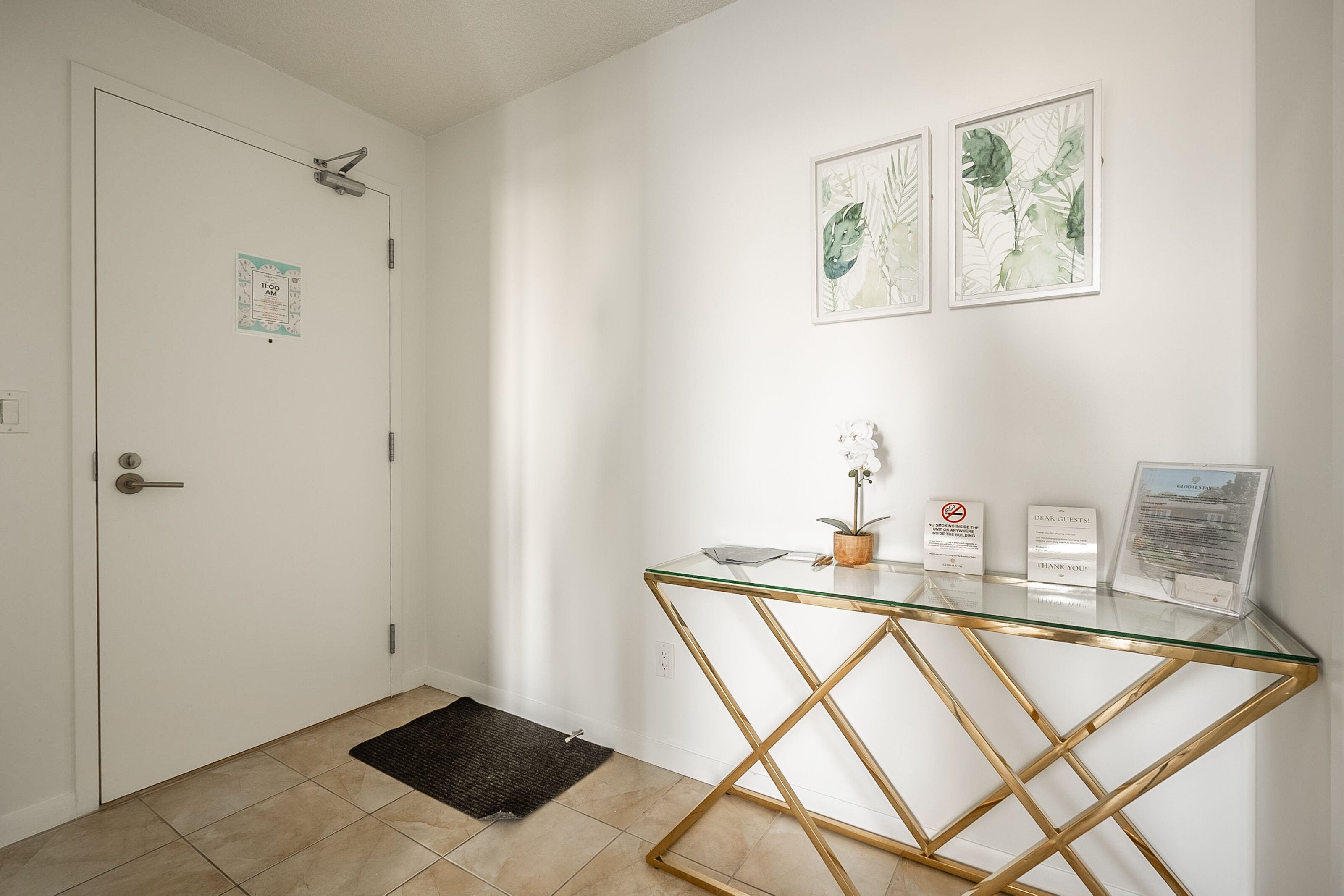 A clean, modern reception area with a glass-top desk, a black doormat, and two framed botanical prints on the wall.