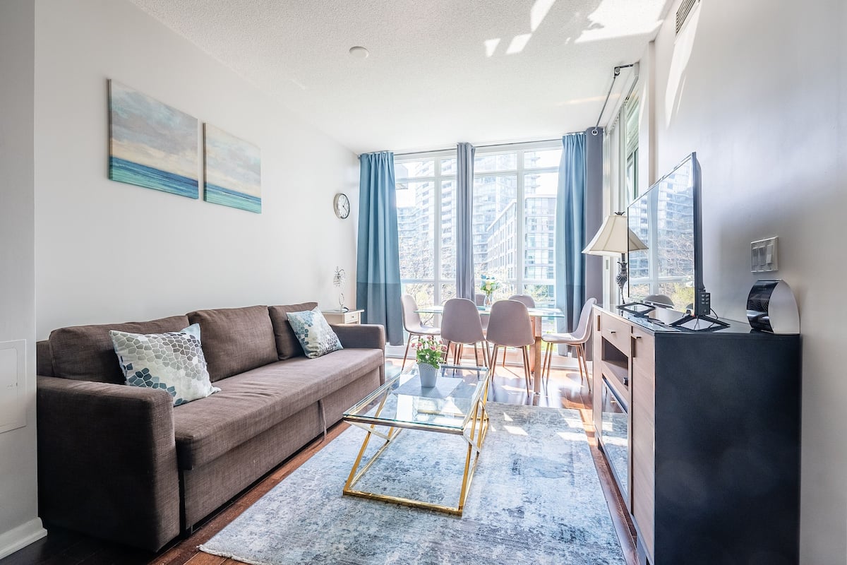 A modern living room with a brown sofa, a glass coffee table, and a view of tall buildings through large windows.