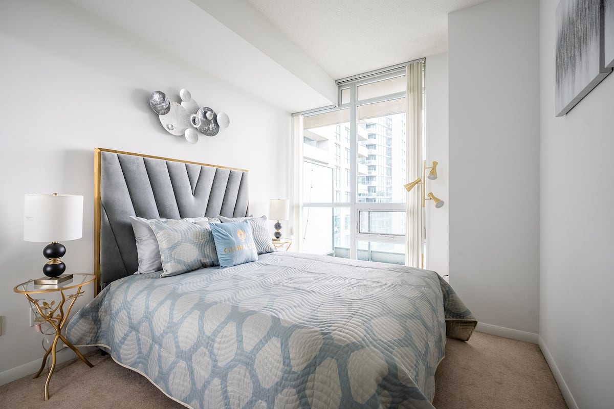 A bedroom with a large bed, a window with a view of buildings, a wall-mounted decorative piece, and a bedside table with a lamp.