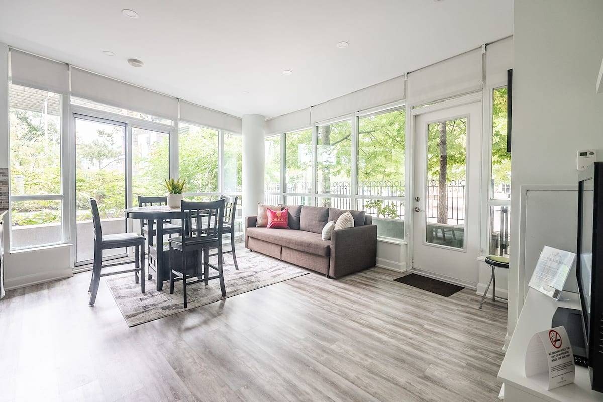 A spacious living area with a sofa, dining table, and chairs, large windows, and a flat-screen TV.