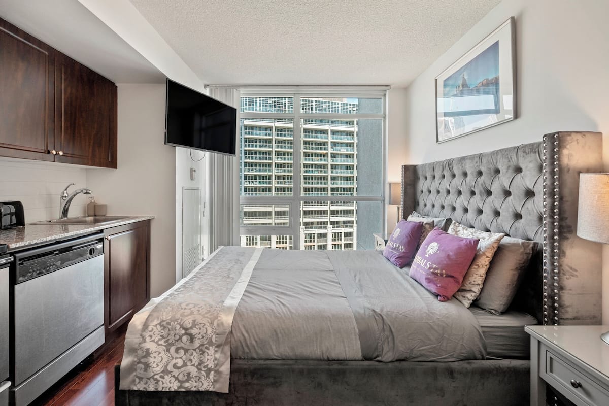 A bedroom with a large bed, a television, a window with a city view, and a kitchen area.