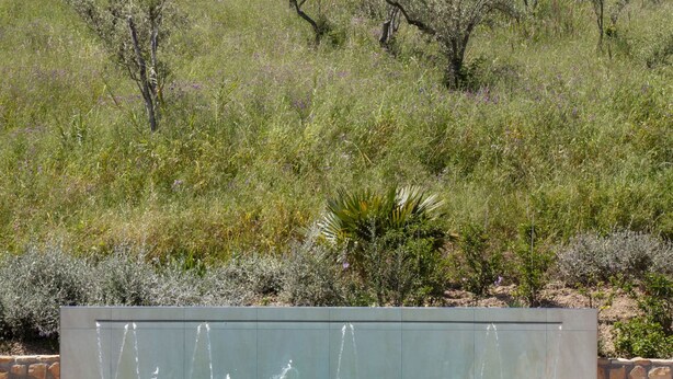 A swimming pool with a water feature, surrounded by a stone wall and a grassy hill with trees. - Villa Cammà (Mirto)