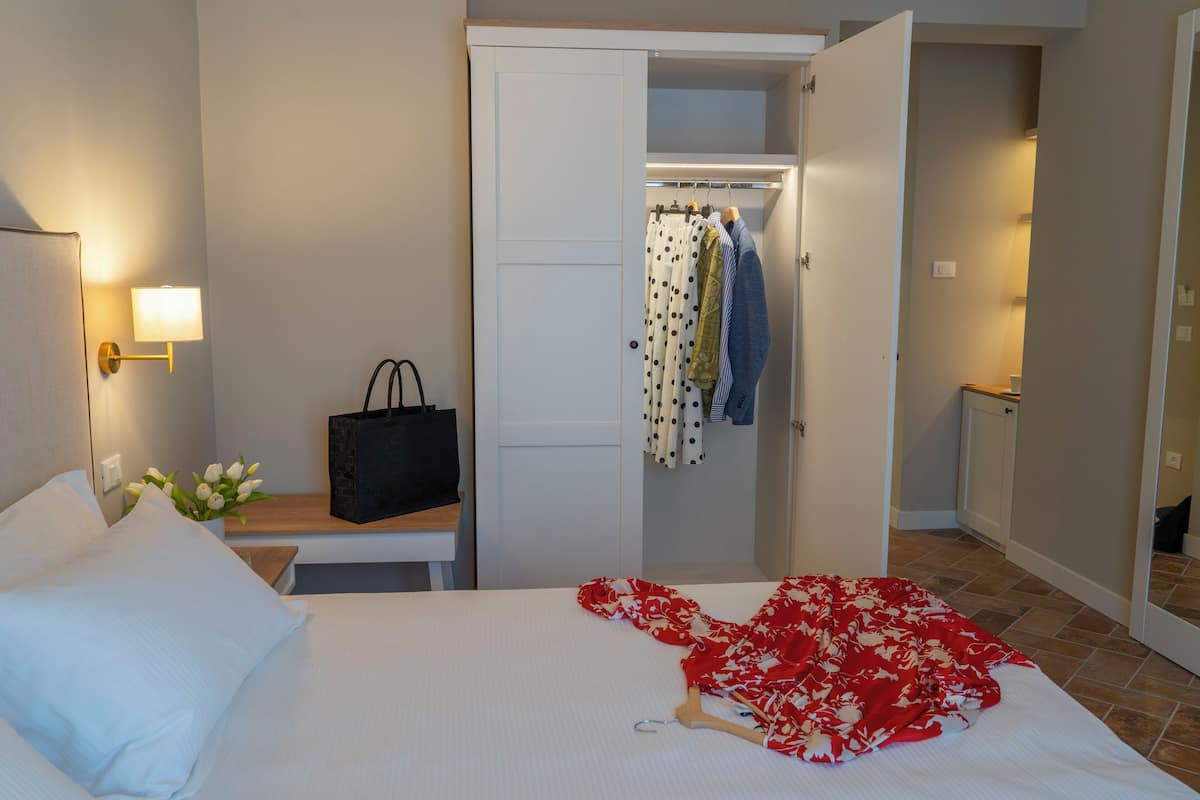 Une chambre d’hôtel avec un lit, une table de chevet avec une lampe, une armoire remplie de vêtements et une salle de bain visible à travers une porte ouverte.
