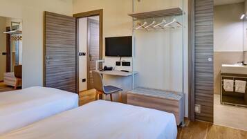 Habitación de hotel con dos camas, un escritorio con silla, televisión y baño con espejo y perchero para toallas.