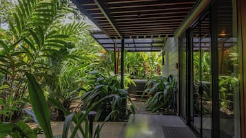 A covered outdoor area with a wooden ceiling, surrounded by lush green plants and a pathway leading to a glass door.