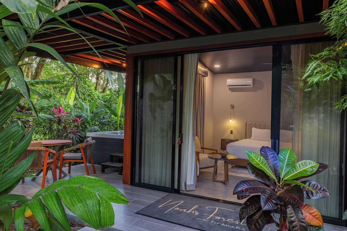 A modern hotel room with a sliding glass door leading to a balcony, a bed, a bedside table, and a small seating area.