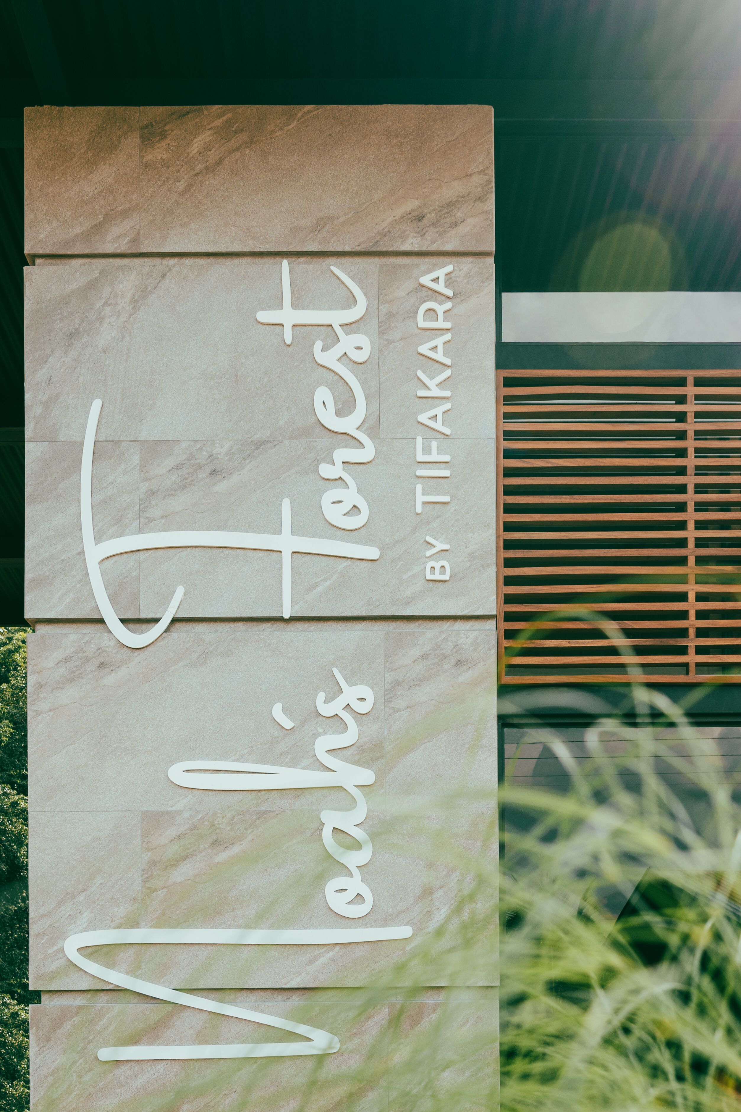 A vertical sign with the text "Forest by TIFAKARA" in a modern font, mounted on a building with a wooden slatted window to the right.