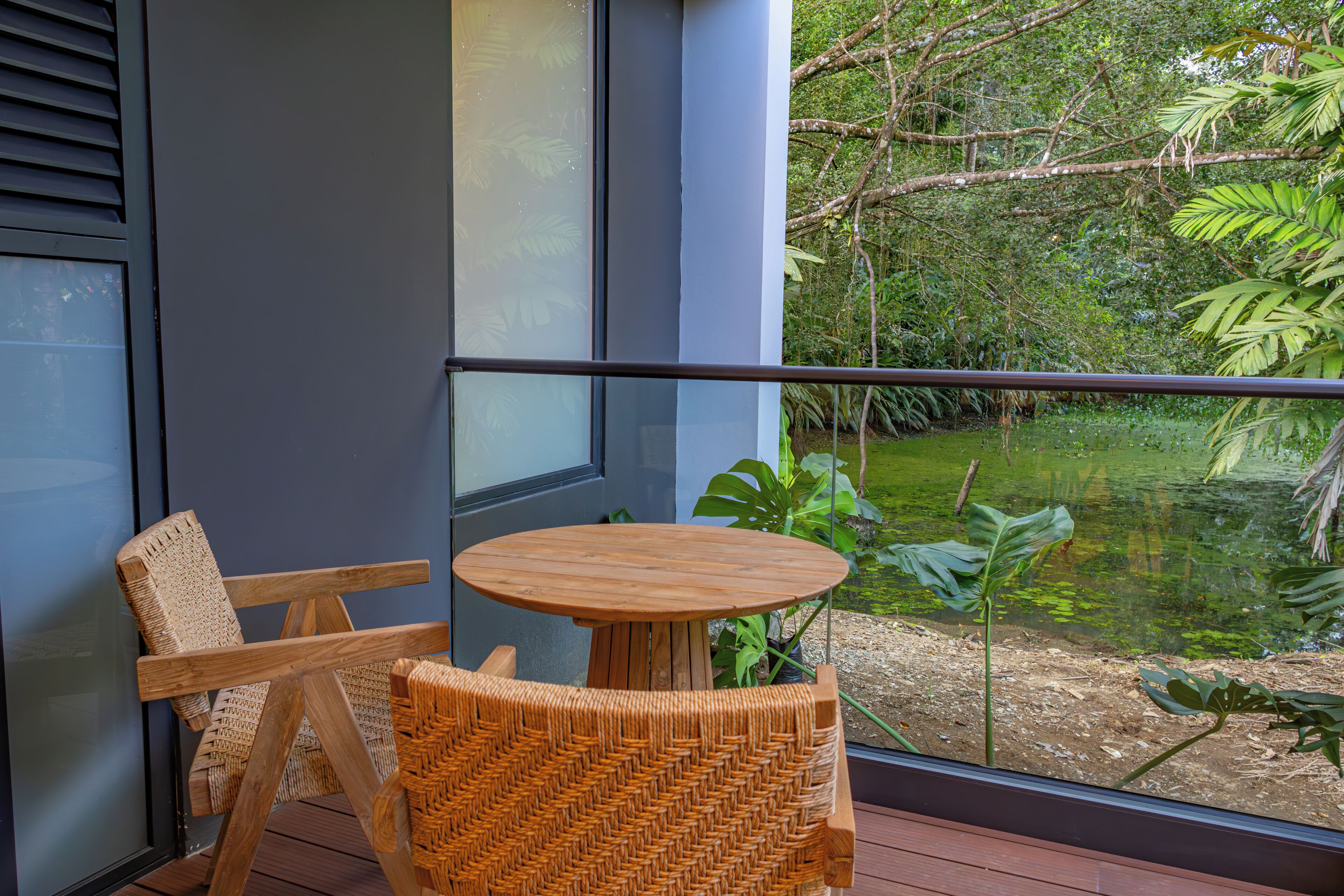 Un balcón con una silla de mimbre y una pequeña mesa de madera, con vista a un estanque rodeado de abundante vegetación.