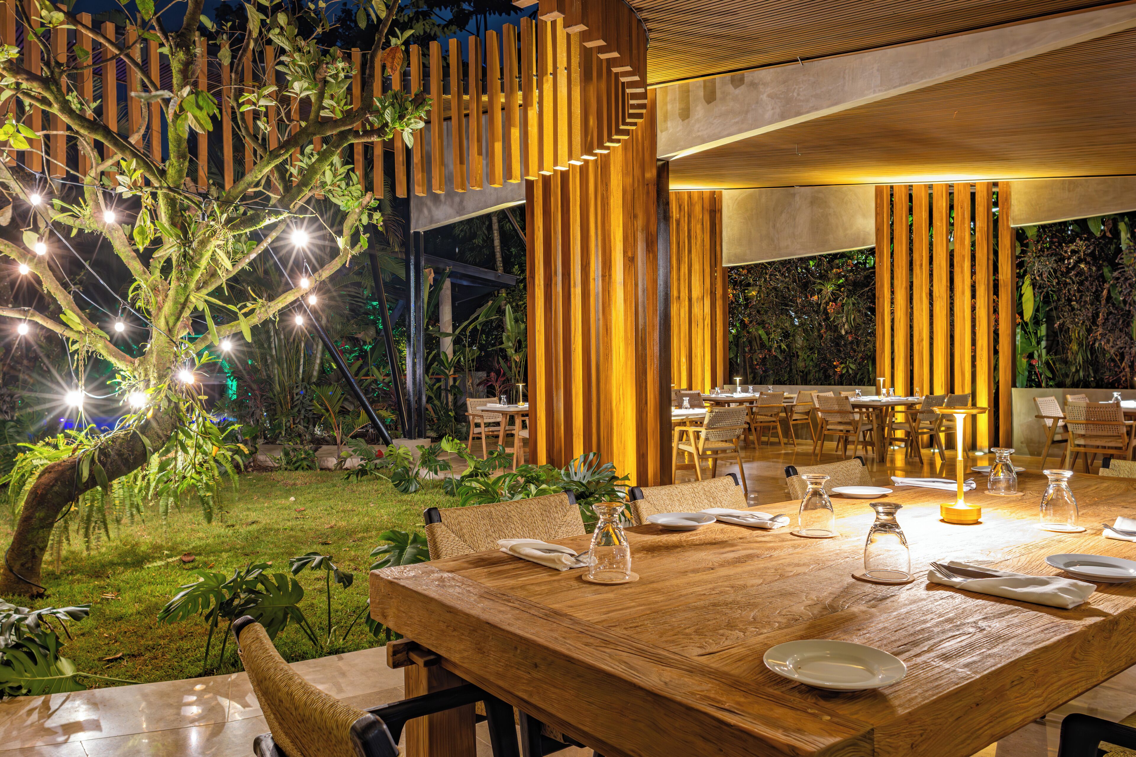 An outdoor dining area with a wooden table set for two, surrounded by chairs, and a tree with string lights.