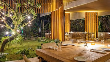 An outdoor dining area with a wooden table set for two, surrounded by chairs, and a tree with string lights.