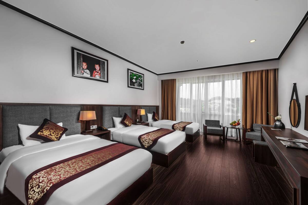 Habitación de hotel con tres camas, un escritorio y una silla. Hay dos cuadros en la pared, una lámpara y un florero con flores.