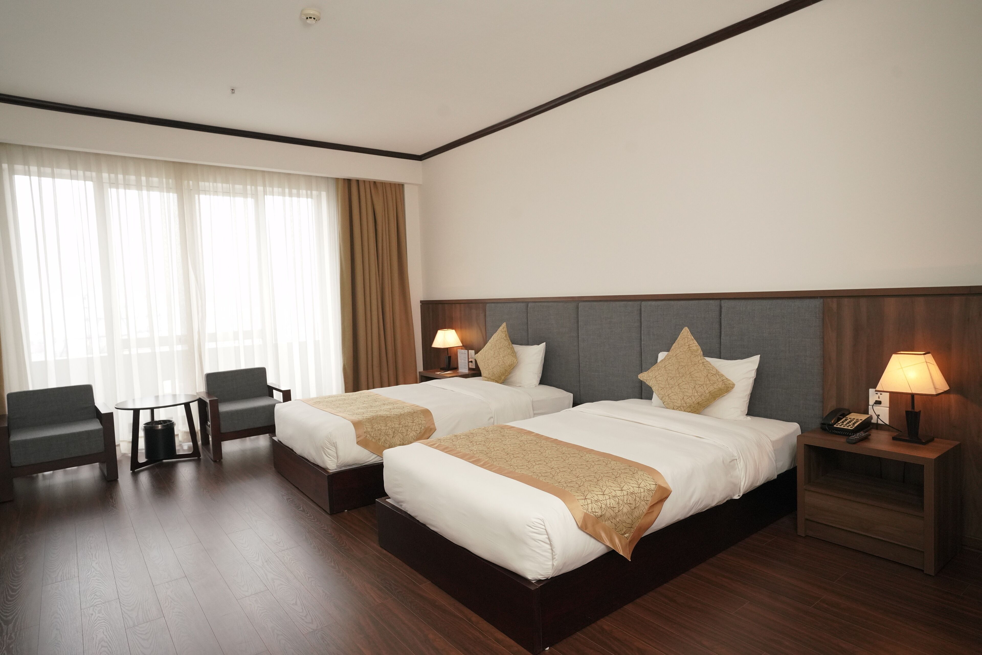 Habitación de hotel con dos camas, un escritorio, sillas y un ventanal grande con cortinas.
