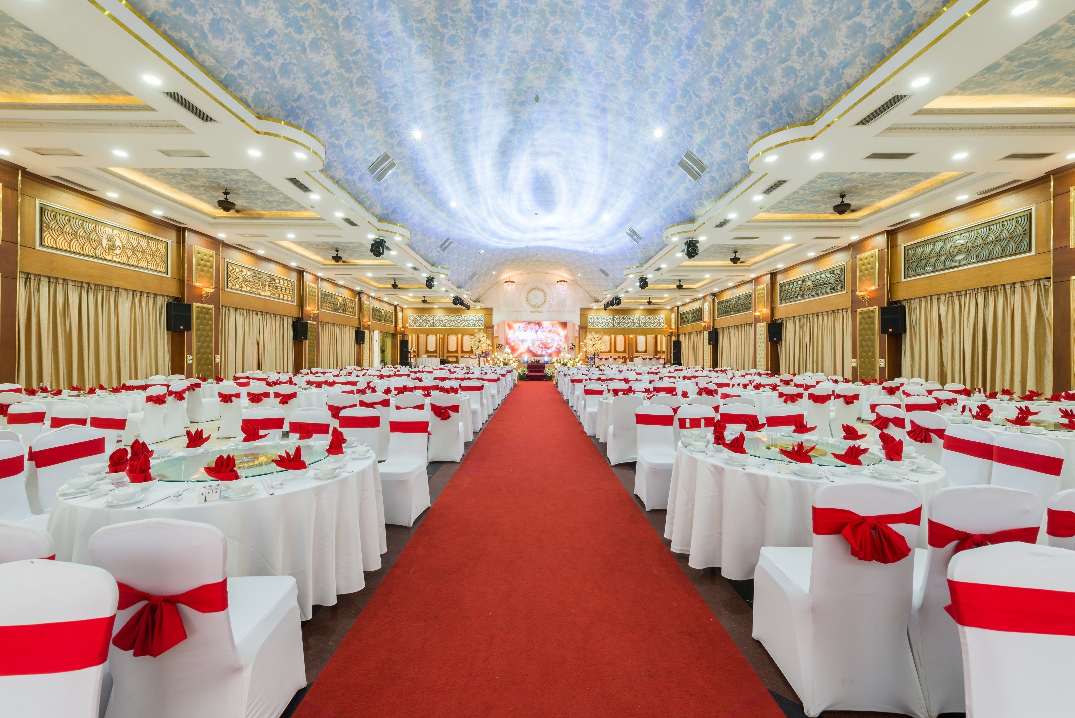 A banquet hall with rows of round tables, each with white tablecloths and red sashes, and a red carpet leading to the front.