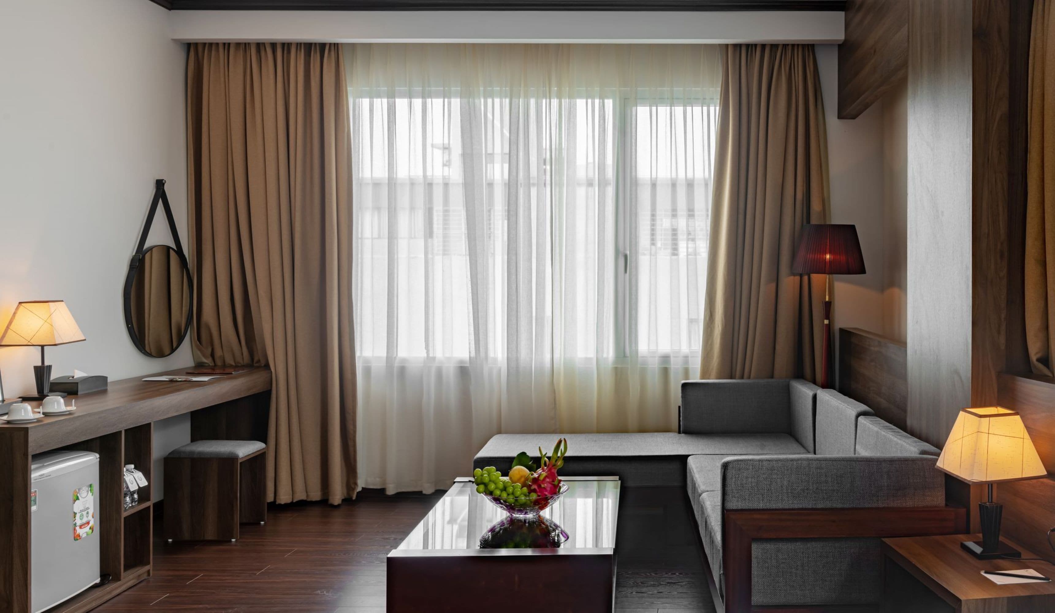 A modern hotel room with a sofa, a coffee table with fruit, a desk with a lamp, and a view of the city through large windows.