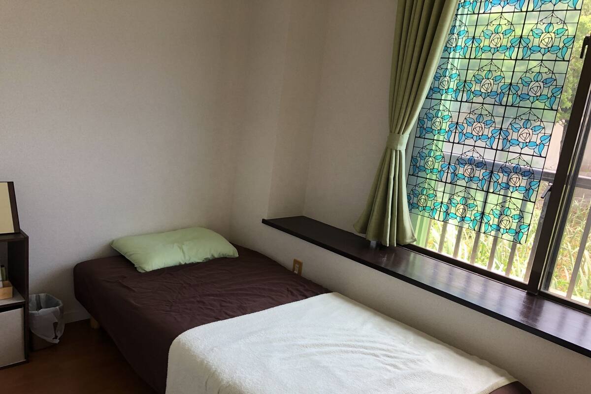 A small, simple room with a bed, a nightstand, a window with a curtain, and a view of greenery outside.