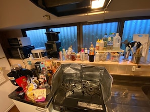 A kitchen with a gas stove, microwave, and coffee maker. There are various bottles and containers on the counter. - Agaitandygama (Miyakojima)