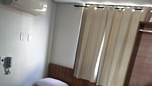 A hotel room with a bed, a blue bedspread, a wooden headboard, a curtain, a wall-mounted air conditioner, and a shelf with towels. - Hotel Campina Executivo (Campina Grande)