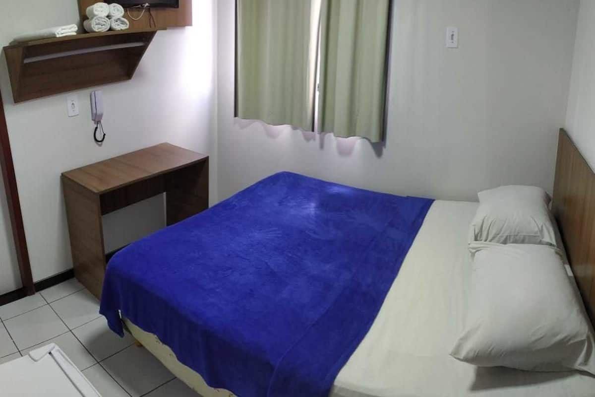 A single bed with a blue blanket, a wooden nightstand, a television above the bed, and a window with curtains.