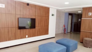 A lobby with a flat-screen TV mounted on a wooden wall, two blue cushioned seating areas, and a fire extinguisher mounted on the wall. - Hotel Campina Executivo (Campina Grande)