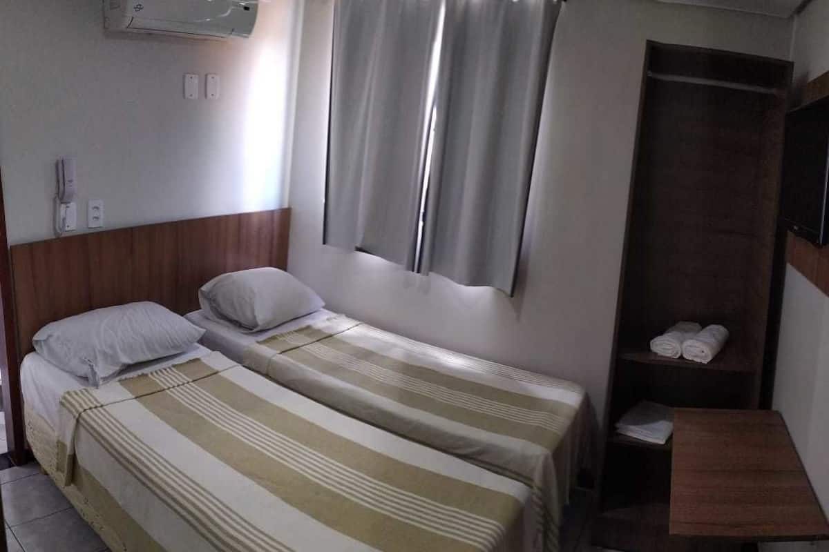 A hotel room with two beds, a wooden headboard, a small wooden shelf with towels, and a window with curtains.