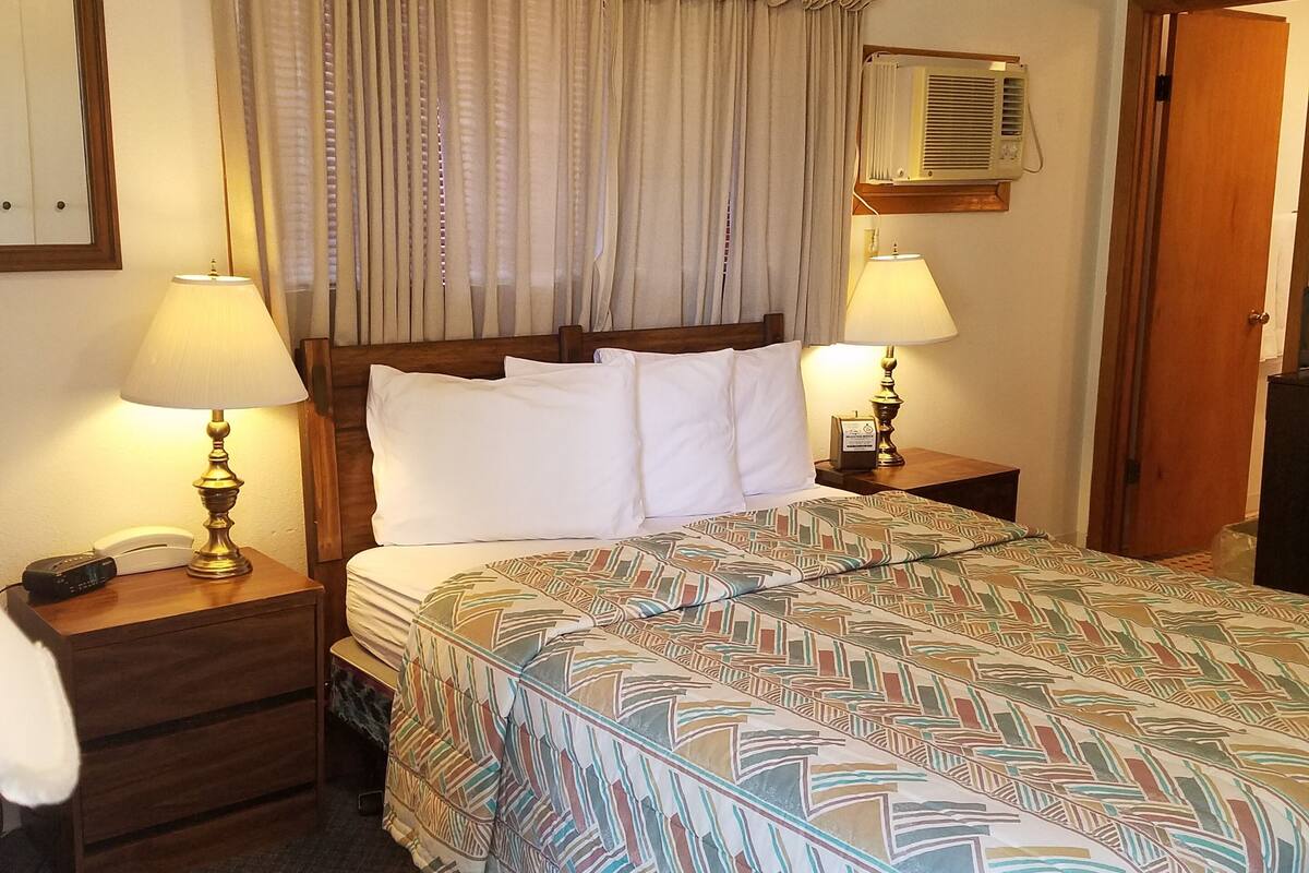 A hotel room with a large bed, two bedside tables with lamps, a wooden headboard, a wall-mounted air conditioner, and a door.