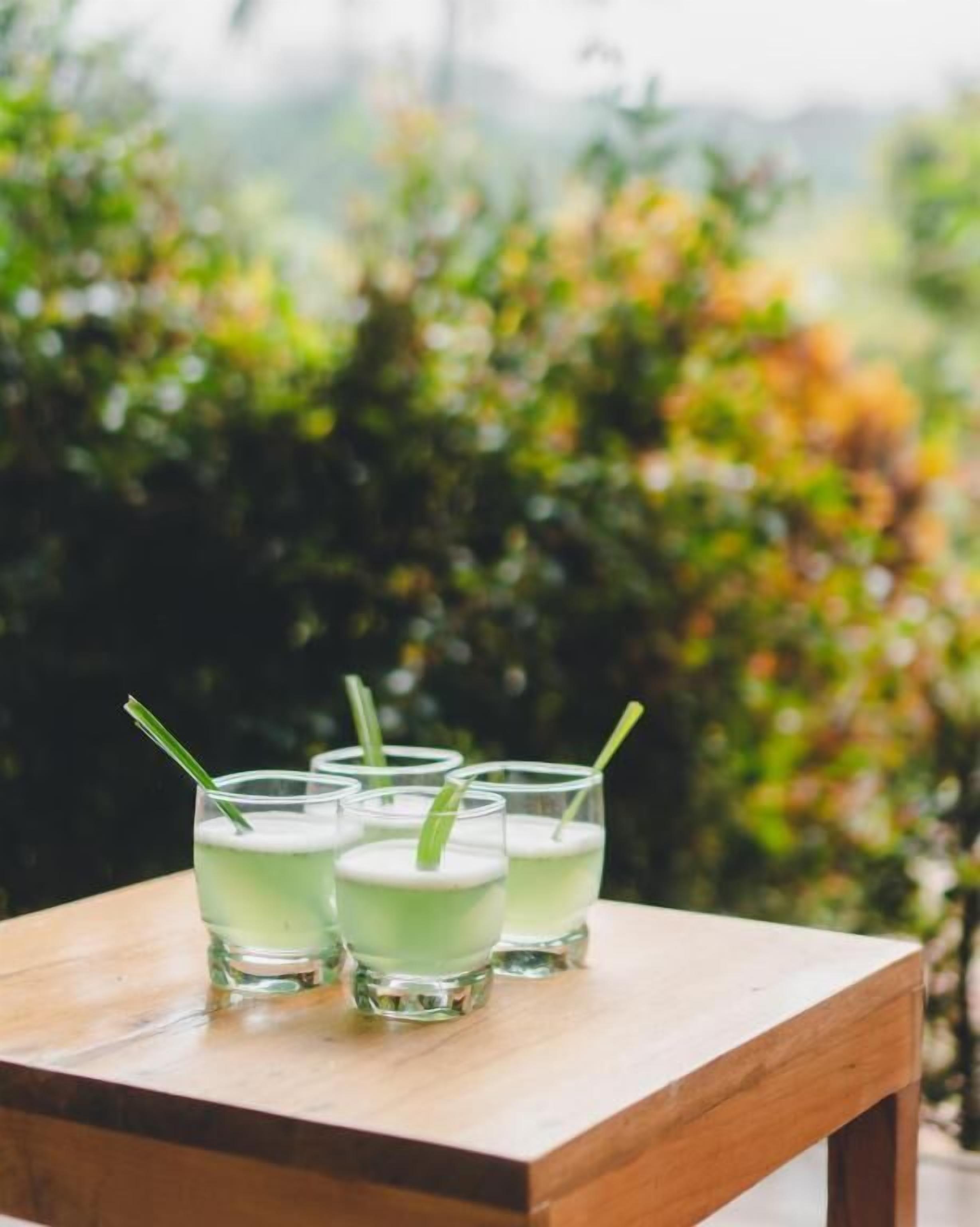 Four glasses of a green beverage on a wooden table with a blurred outdoor background.