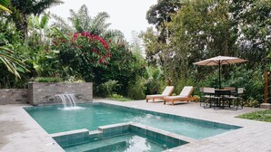 A rectangular swimming pool with a waterfall feature, surrounded by lush greenery and a seating area with a table and chairs.