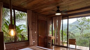 A bedroom with a bed, wooden ceiling, large windows, and a view of greenery.