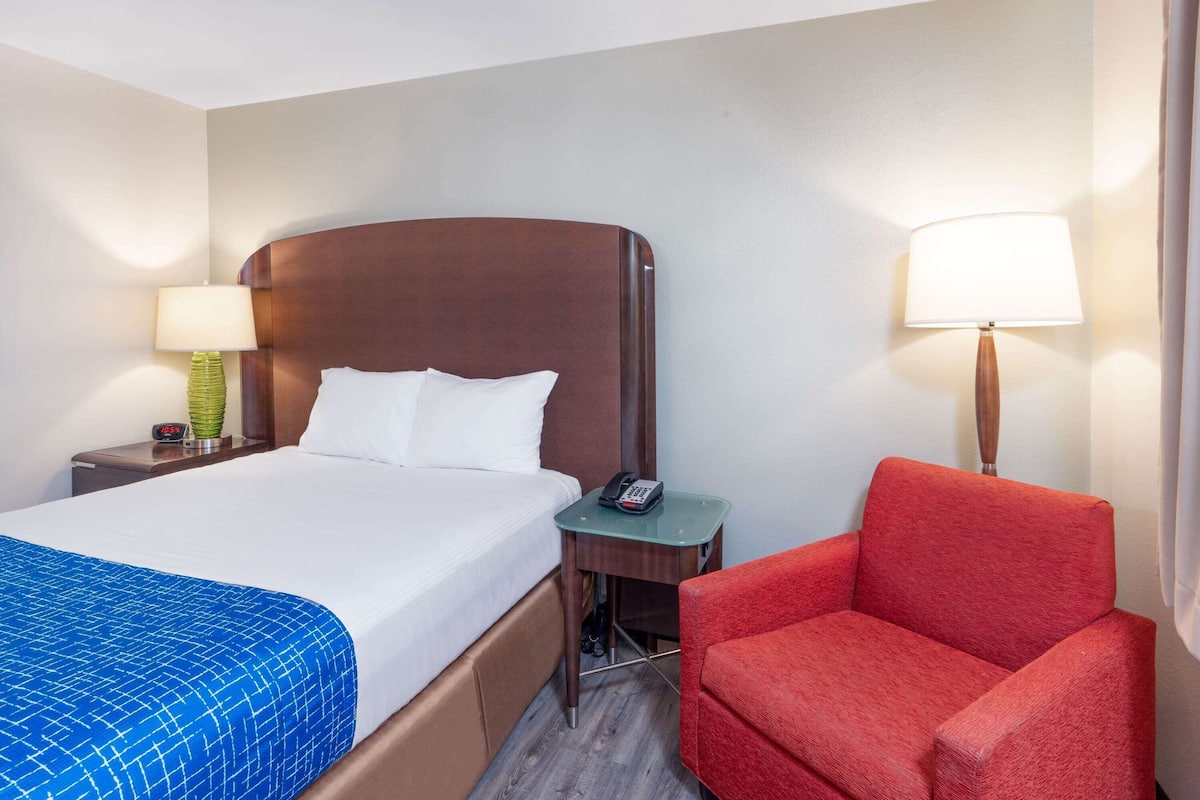 A hotel room with a bed, a red armchair, a nightstand with a lamp, and a small table with a phone.