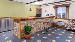 Un área de recepción con mostrador de madera, pared de piedra, planta en maceta, taburete de bar, sofá y ventana con cortinas.