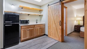 Cocina compacta con refrigerador negro, microondas y armarios de madera. Una puerta corrediza tipo granero da acceso a un dormitorio con cama y mesita de noche.