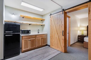 Cozinha compacta com geladeira preta, micro-ondas e armários de madeira. Uma porta corrediça tipo celeiro dá acesso a um quarto com cama e mesa de cabeceira.