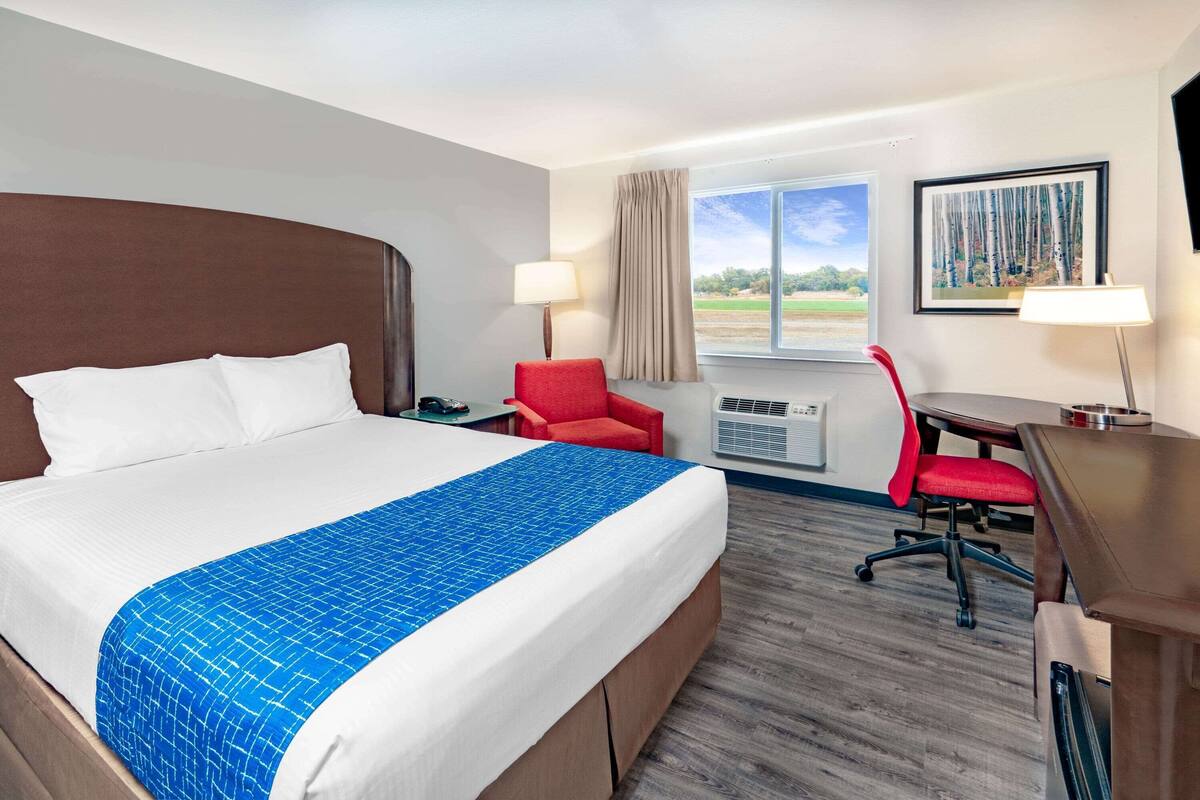 A hotel room with a large bed, a desk, a red chair, a window with a view, and an air conditioning unit.