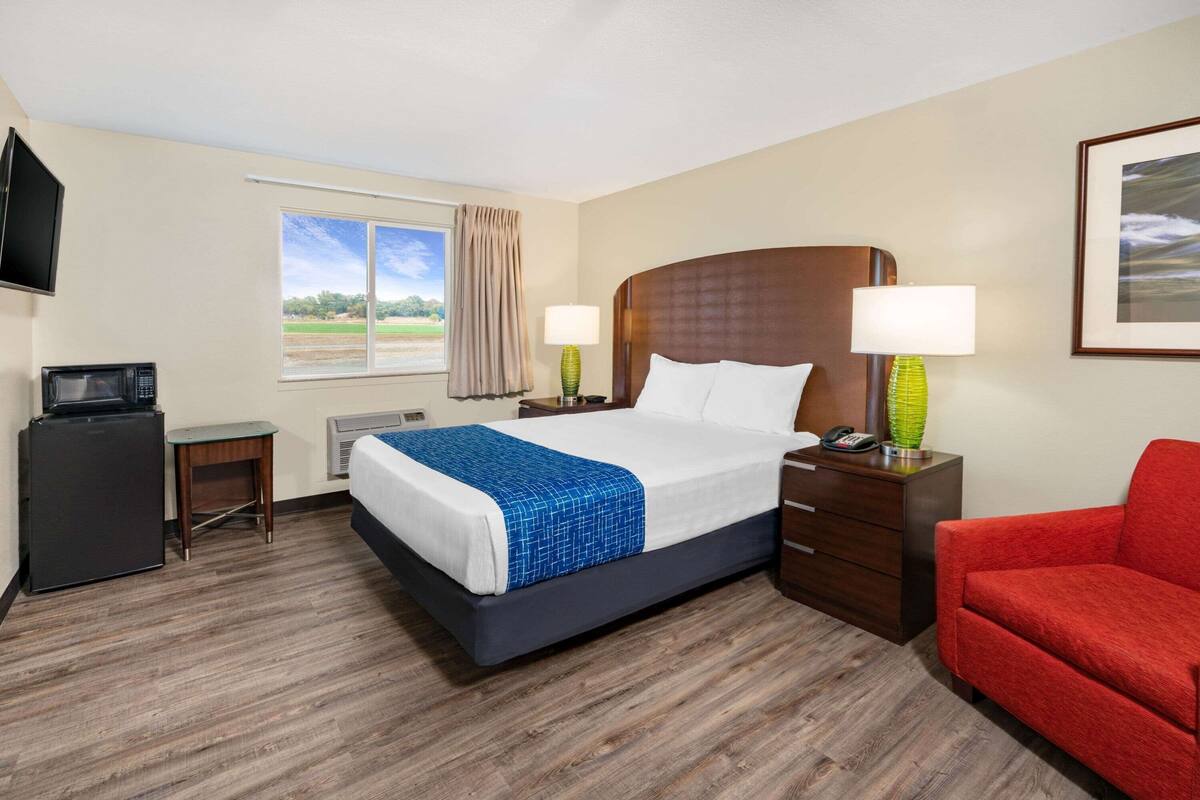 A hotel room with a bed, bedside lamps, a TV, a small desk with a microwave, and a red chair.
