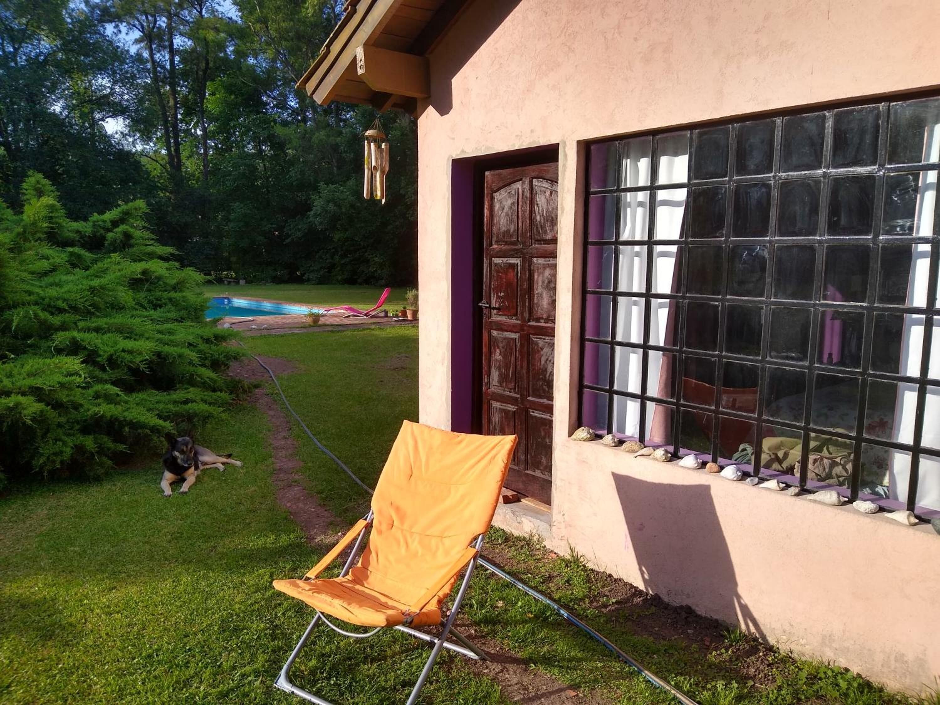 Una casa con puerta de madera, ventana con diseño de cuadrícula y césped con un perro y una silla.