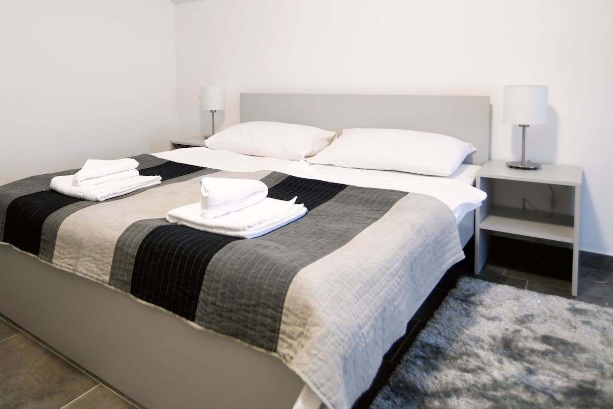A neatly made bed with a grey and black striped bedspread, white pillows, and folded white towels on the bed. There is a white bedside table with a lamp on it.
