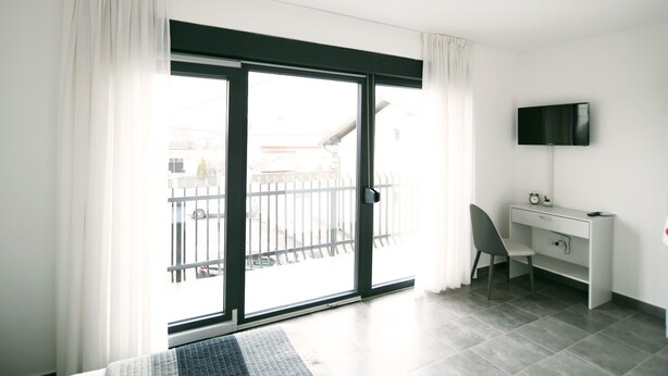 A modern hotel room with a sliding glass door to a balcony, a desk with a chair, a flat-screen TV mounted on the wall, and a small clock on the desk.