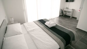 A neatly made bed with white linens, a grey and black striped blanket, and a folded towel on the bed. There is a white bedside table with a lamp, a small clock, and a power outlet. A desk with a chair is visible in the background, and a TV is mounted on the wall.