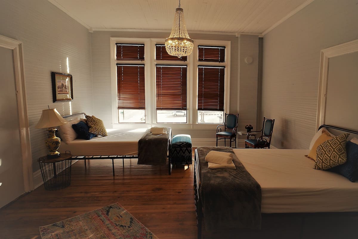 Quarto com uma cama grande, duas cadeiras, uma mesa pequena, um abajur e uma janela com persianas.