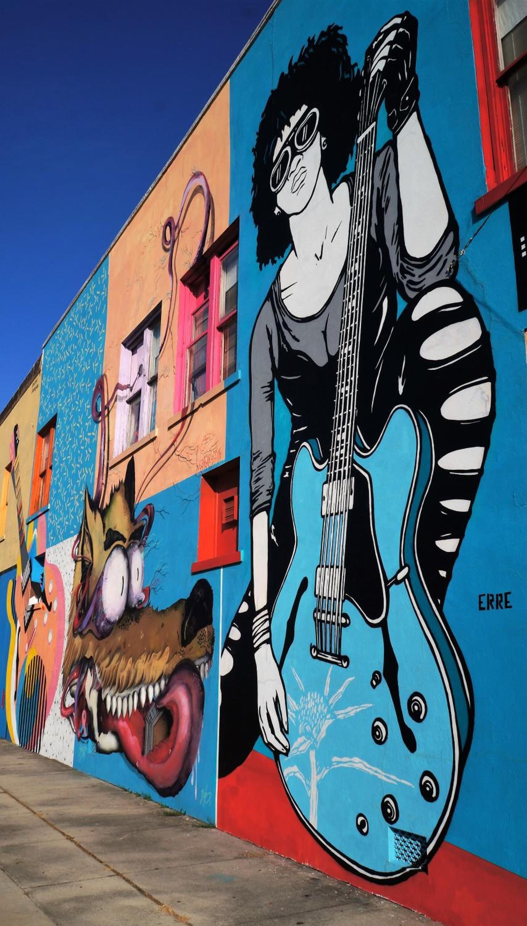 Um mural colorido na fachada de um edifício retrata uma pessoa tocando violão e um animal estilizado.