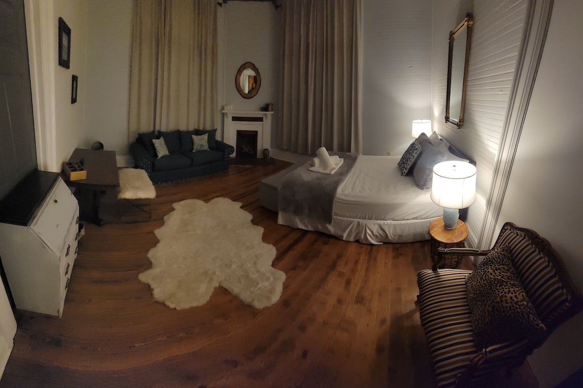 Quarto de hotel acolhedor com cama, sofá, mesa pequena com abajur, lareira e tapete grande.