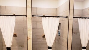 Zona de ducha con tres cortinas de ducha blancas sujetas a una barra central, paredes de azulejos color beige y suelo de azulejos.