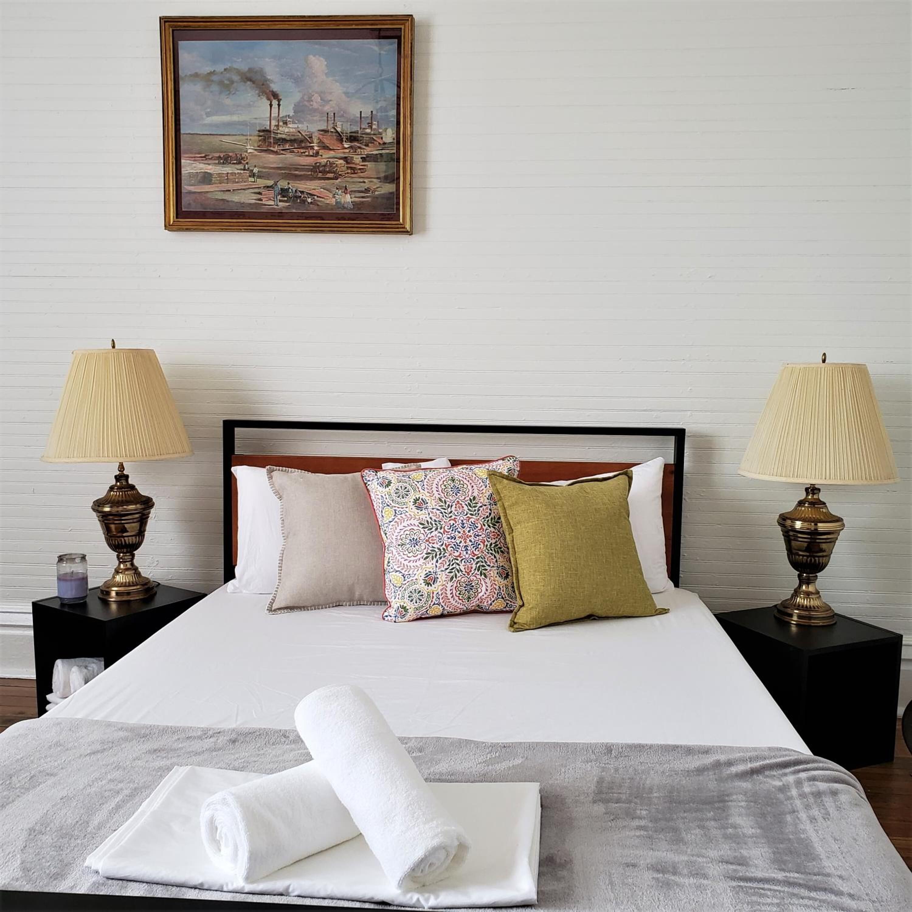 A neatly made bed with a white duvet, two decorative lamps, and a painting above it.