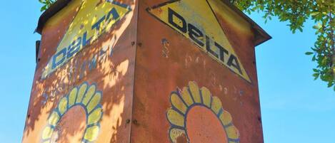 A rusted, weathered Delta sign with a sun design, mounted on a pole in an urban setting with buildings in the background.