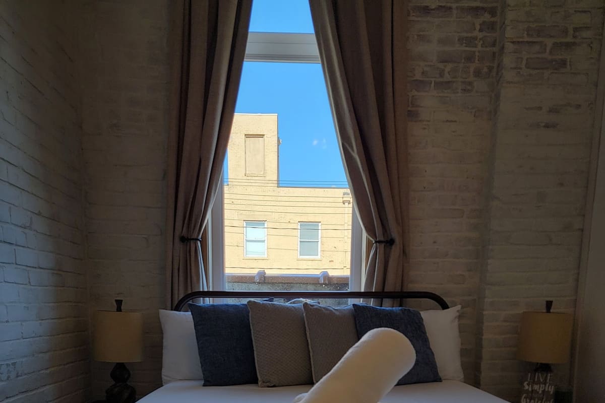 Cama arrumada com uma toalha enrolada, duas luminárias de cabeceira e vista de um edifício através de uma janela com cortinas.