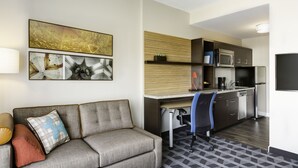 A modern hotel room with a grey sofa, a desk with a chair, a kitchen area with a microwave and refrigerator, and artwork on the wall. - TownePlace Suites by Marriott Medicine Hat (Medicine Hat)
