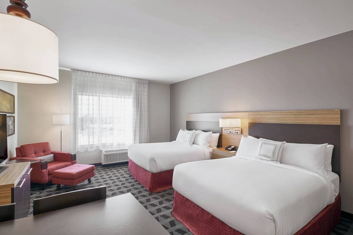 Ein Hotelzimmer mit zwei Betten, einem Schreibtisch, einem Stuhl und einem großen Fenster mit Vorhängen.