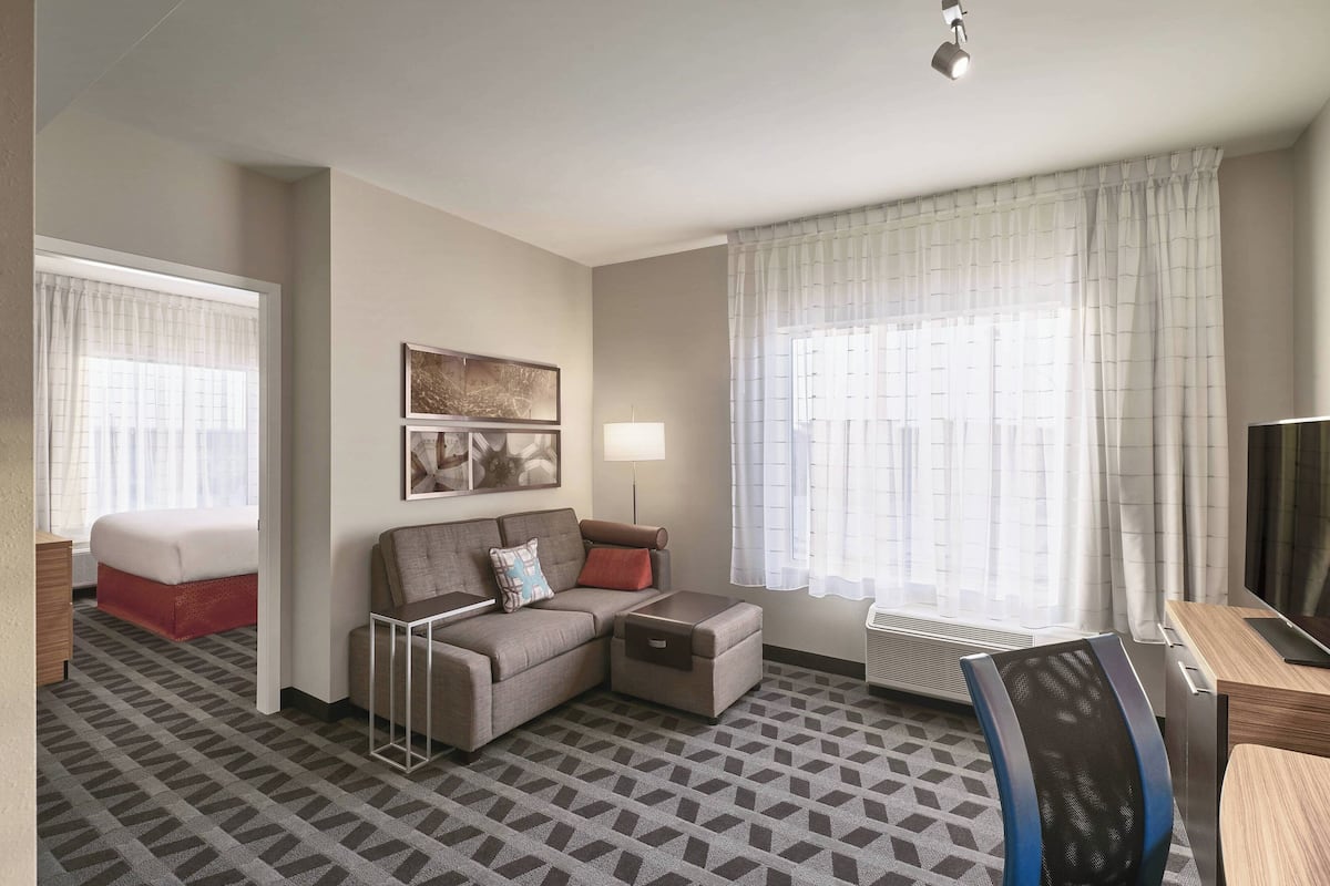 Ein Hotelzimmer mit einer Couch, einem kleinen Tisch, einem Sessel, einem Bett, einem Fernseher und einem Fenster mit Vorhängen.