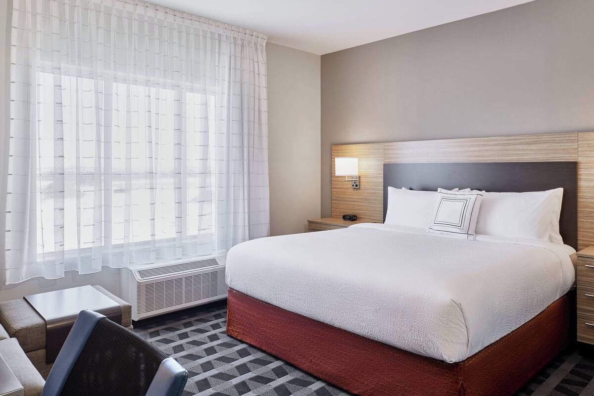 Ein Hotelzimmer mit einem großen Bett, einem Nachttisch, einem Sessel und einem Fenster mit durchsichtigen Vorhängen.