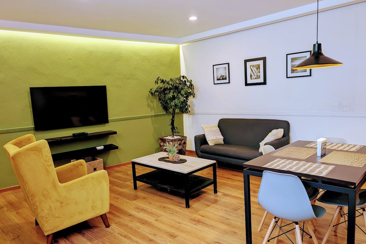 Una sala moderna con una televisión de pantalla plana, un sofá, una mesa de comedor con sillas y una planta en maceta.